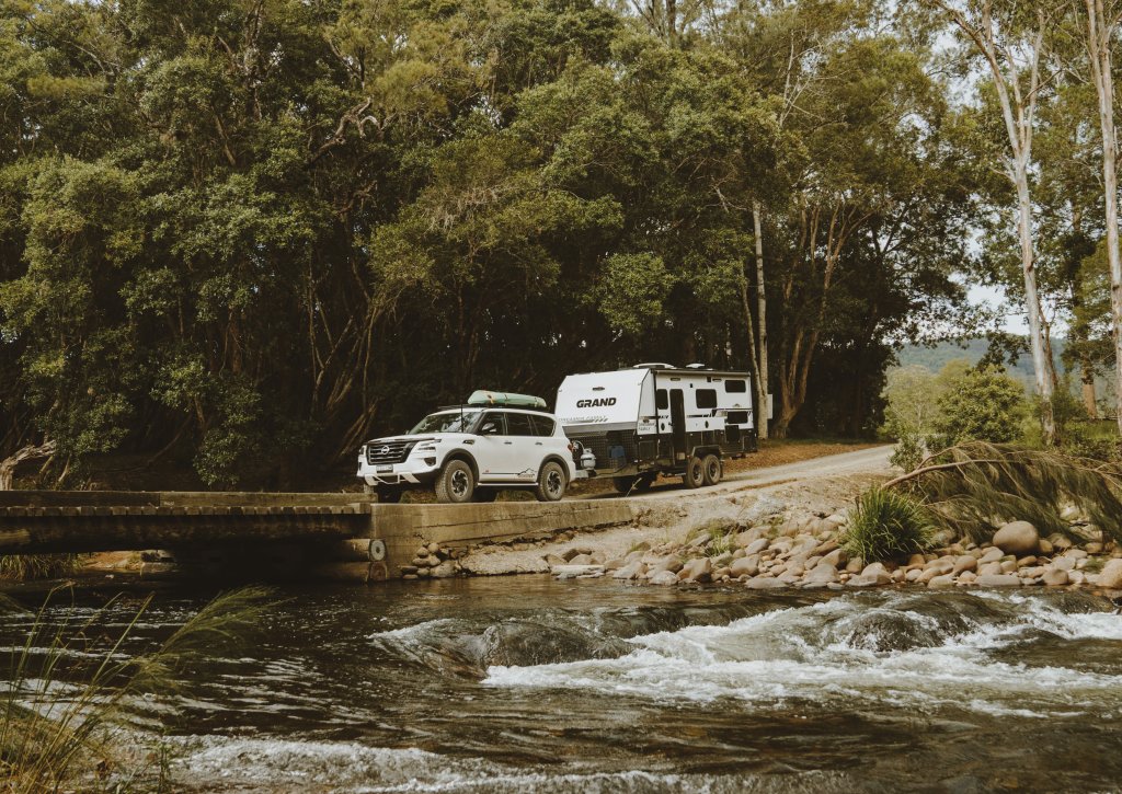 A white car towing a Grand caravan over a bridge over a river in a green forest in Australia